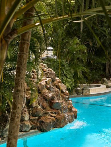 A rooftop infinity pool overlooking a cityscape is surrounded by lounge chairs, with a clear blue sky and scattered clouds above.