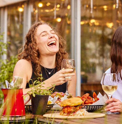 Guests enjoy drinks and laughter on a sunlit patio, surrounded by greenery at a lively hotel bar.