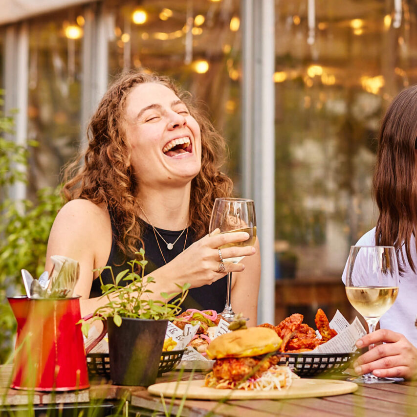 Guests enjoy drinks and laughter on a sunlit patio, surrounded by greenery at a lively hotel bar.