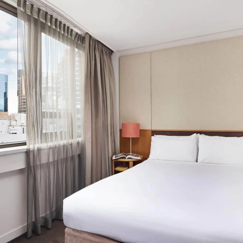 Bright, modern hotel room with city view, featuring a comfortable double bed, bedside lamps, and large window with sheer curtains.
