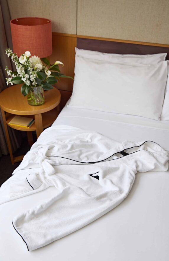 Cosy hotel room with plush bed, soft white robe, bedside table, flower arrangement, and ambient lighting for relaxation.
