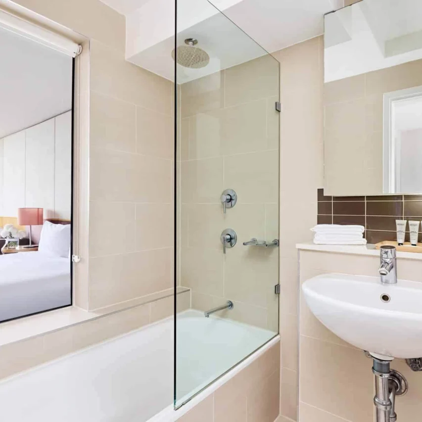 Modern hotel bathroom with glass shower, cosy bed view, plush towels, luxury toiletries, inviting stay, bright natural light.