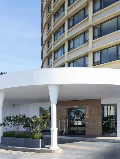 Modern hotel entrance with elegant curved canopy, large windows, and welcoming garden, promising a luxurious stay.