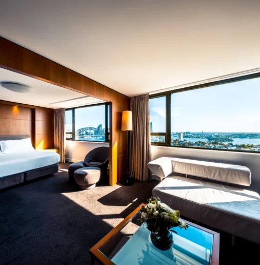 Spacious hotel suite with Harbour view, plush bed, modern furnishings, large windows, and cosy seating. Perfect relaxation spot.