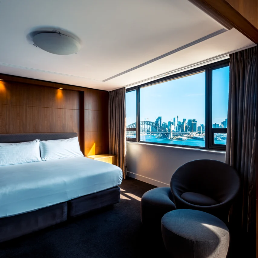 Luxurious hotel room with city skyline view, plush king-size bed, modern decor, and large window.