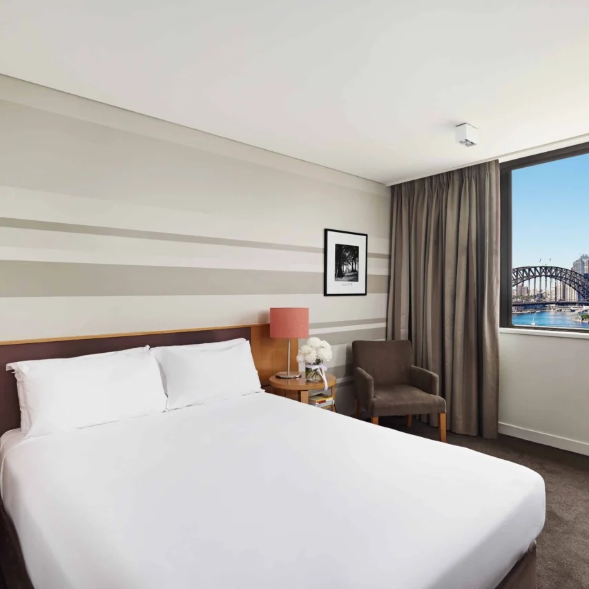 Modern hotel room with plush bed, city view, and Harbour Bridge, ideal for a comfortable stay and scenic experience.