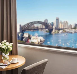 Spacious hotel room with cityscape view, featuring harbour, iconic bridge, fresh flowers, and stylish seating.