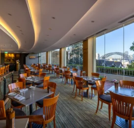 Elegant hotel restaurant with Sydney Harbour Bridge view, wooden chairs, well-set tables, cosy ambiance. Perfect dining spot.