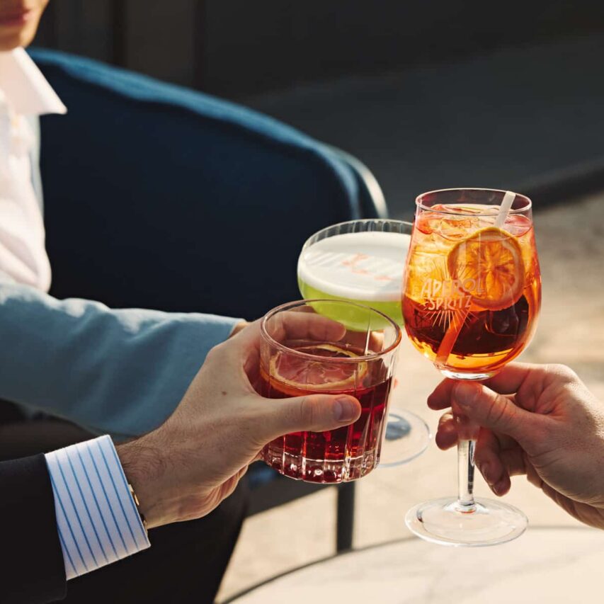 Group of people enjoying a mixed indoor-outdoor restaurant space with green plants, wooden accents, and natural lighting, creating a lively and inviting dining atmosphere.