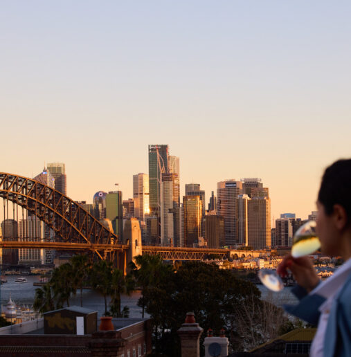 Woman drinking champagne looking out at harbour bridge at sunset
