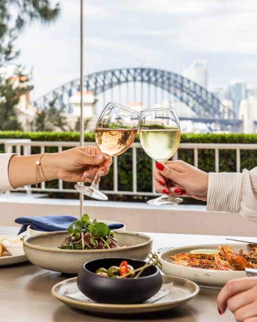 Two people cheers-ing wine over a meal, with the Sydney Harbour Bridge in the background.