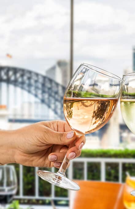 Two wine glasses cheersing in a restaurant with the harbour bridge in the background.