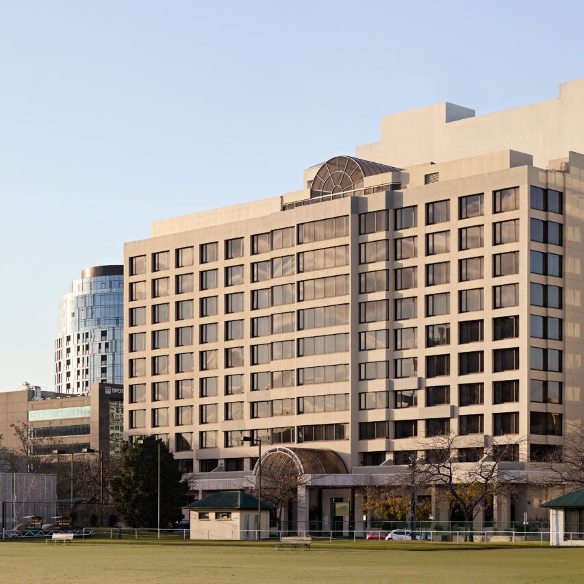 A modern multi-story hotel with a striking glass facade against a blue sky, surrounded by trees and parked cars. The building features angular architectural elements and a spacious, inviting entrance.