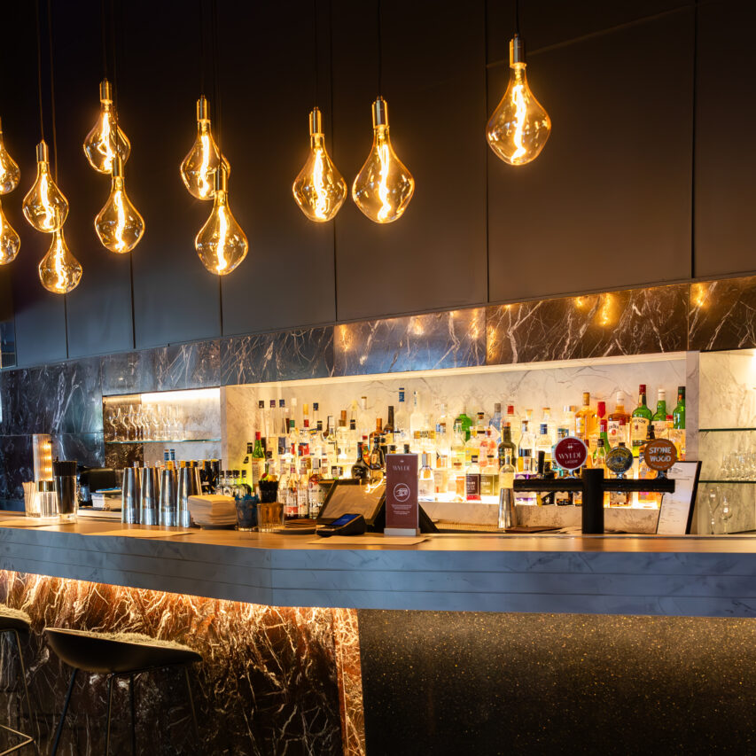 Image of Wylde bar with filament lights overhanging the bar and under bar lighting. there is a wall of spirits and they are also lit up.