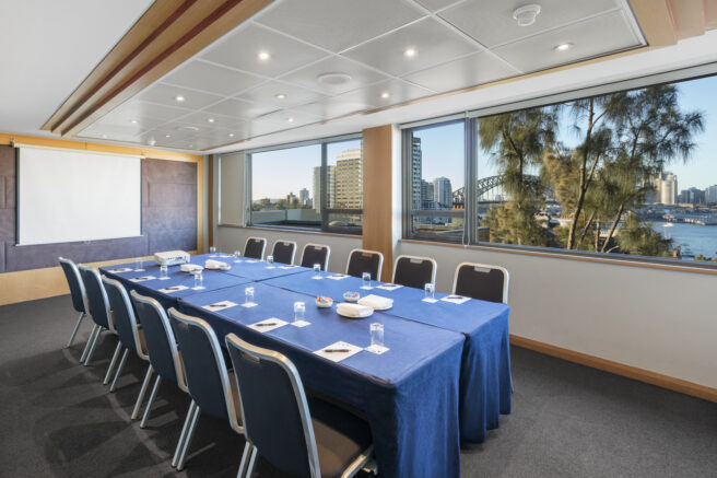 A modern boardroom with a long table and ergonomic chairs, large windows with a view of Sydney Harbour, featuring the iconic Sydney Opera House and Sydney Harbour Bridge.