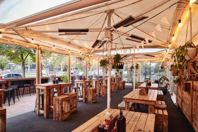 Outdoor restaurant seating area at night with string lights, surrounded by greenery and trees, creating a cozy and inviting ambiance. White chairs and tables are neatly arranged.