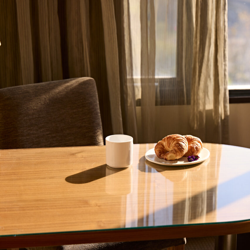 A modern hotel room with a large bed, a sleek armchair, and a cityscape view through a floor-to-ceiling window.