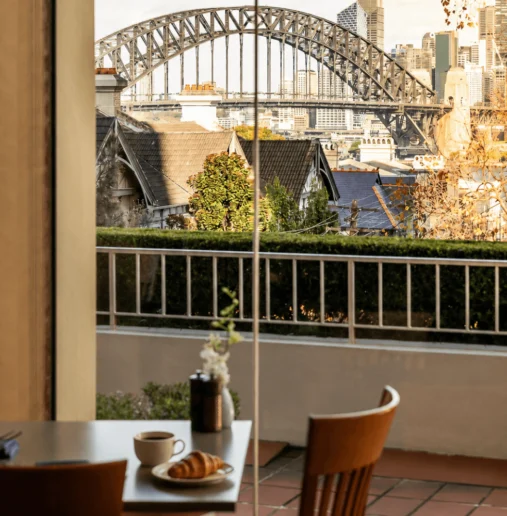 A buffet breakfast spread featuring various pastries, fruits, cereals, and drinks displayed on a marble countertop, with a view of a city skyline through large windows in the background.